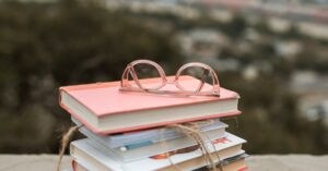 Stack of colorful books with glasses on top, set outdoors with blurred background.