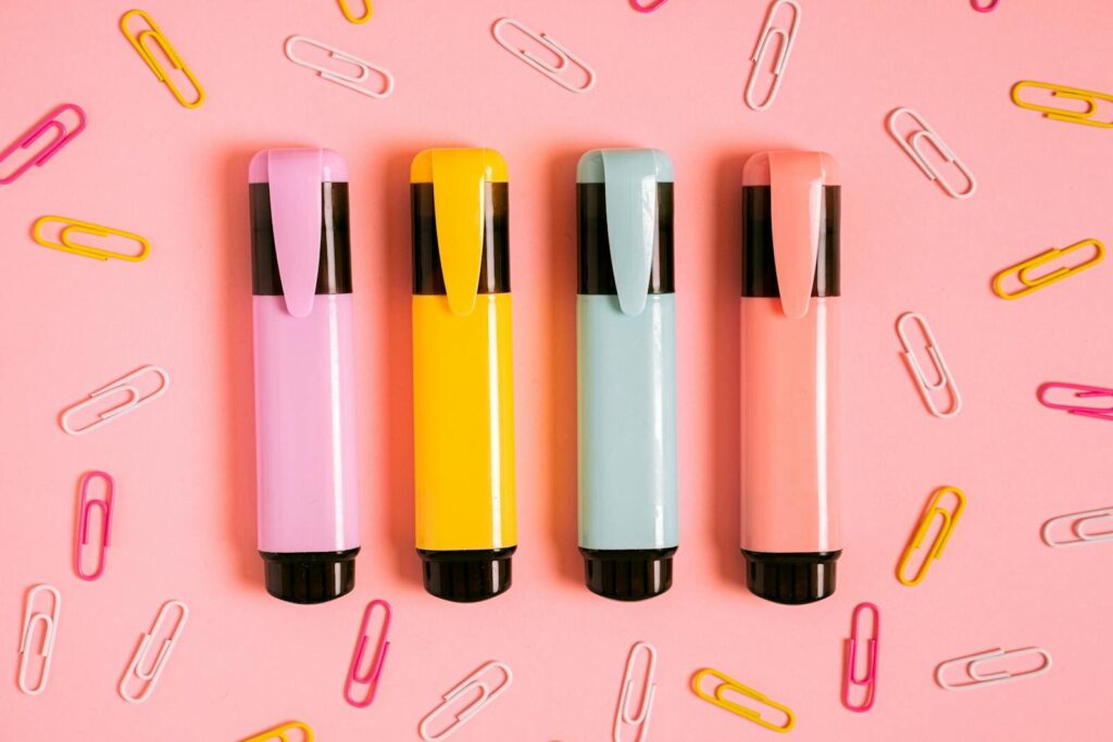 A flat lay of colorful markers and paper clips on a pink surface, perfect for stationery themes.