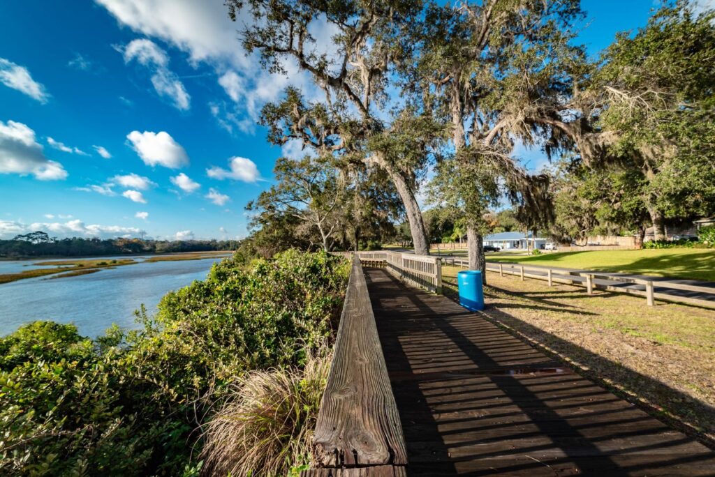 A picturesque boardwalk along a riverbank in Florida on a sunny day, surrounded by lush greenery and blue skies.